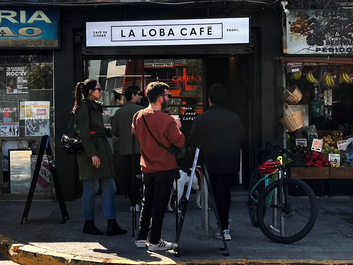 Fachada de La Loba Café: letrero blanco con el nombre, vitrina oscura con el neón Cafeína y personas esperando en la vereda